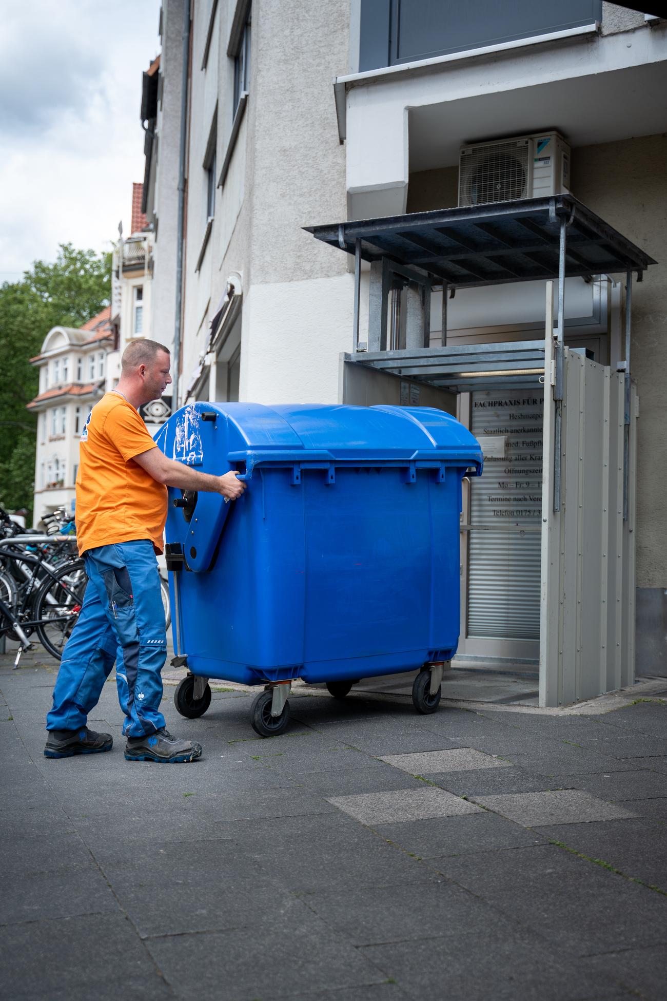 Mitarbeiter im Firmenshirt schiebt einen großen Müllcontainer vor einem Wohngebäude – Koordiniert durch Professionelle Hausverwaltung