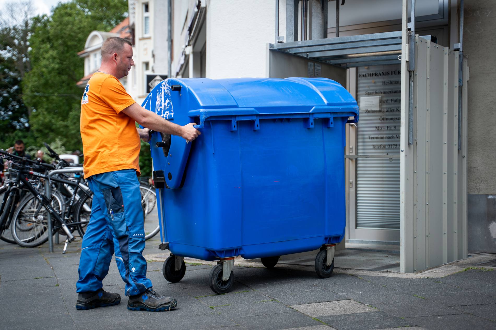 Mitarbeiter im Firmenshirt schiebt einen großen blauen Müllcontainer vor einem Gebäude – Facility Management Maßnahme der professionellen Hausverwaltung